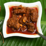 pickling beef - Kerala beef pickle in a white bowl on banana leaf with dried red chilies and spicy oil